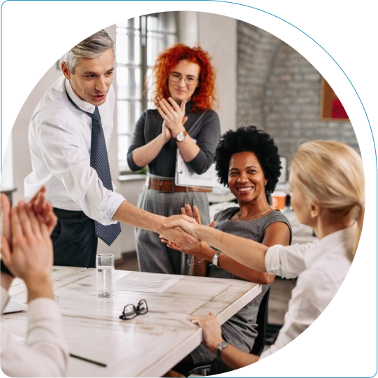 Man and woman shaking hands across a table, while the rest of the group around the desk is clapping their hands cheerfully